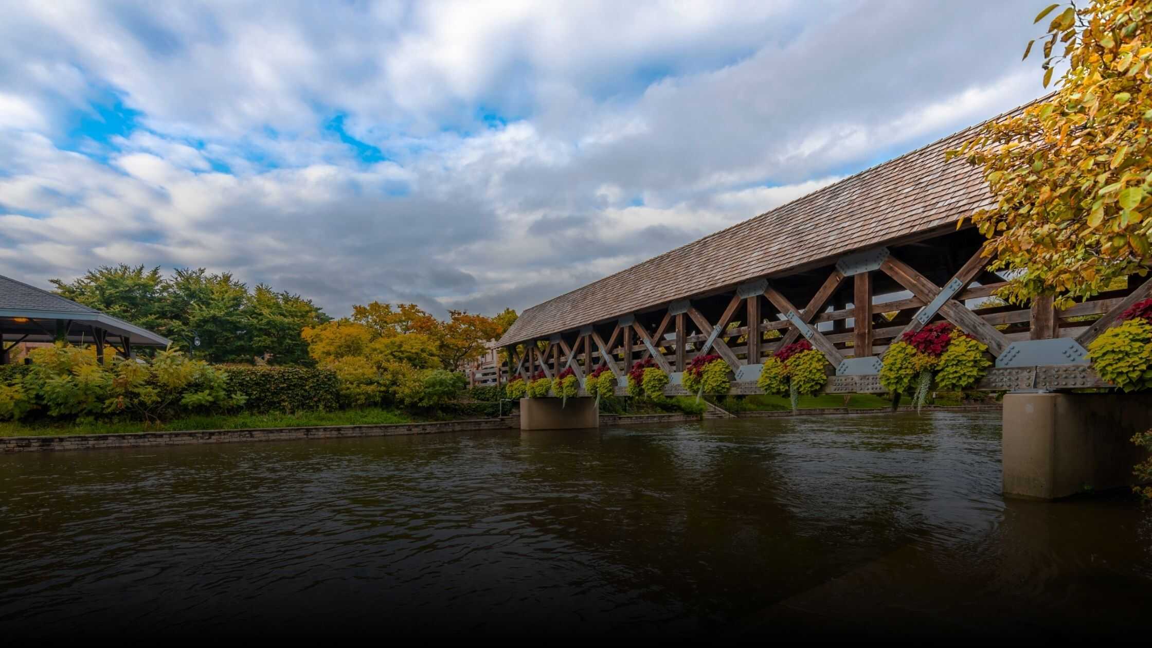 Scenic view of the covered bridge along Naperville's Riverwalk, surrounded by lush greenery, colorful flowers, and partly cloudy skies.